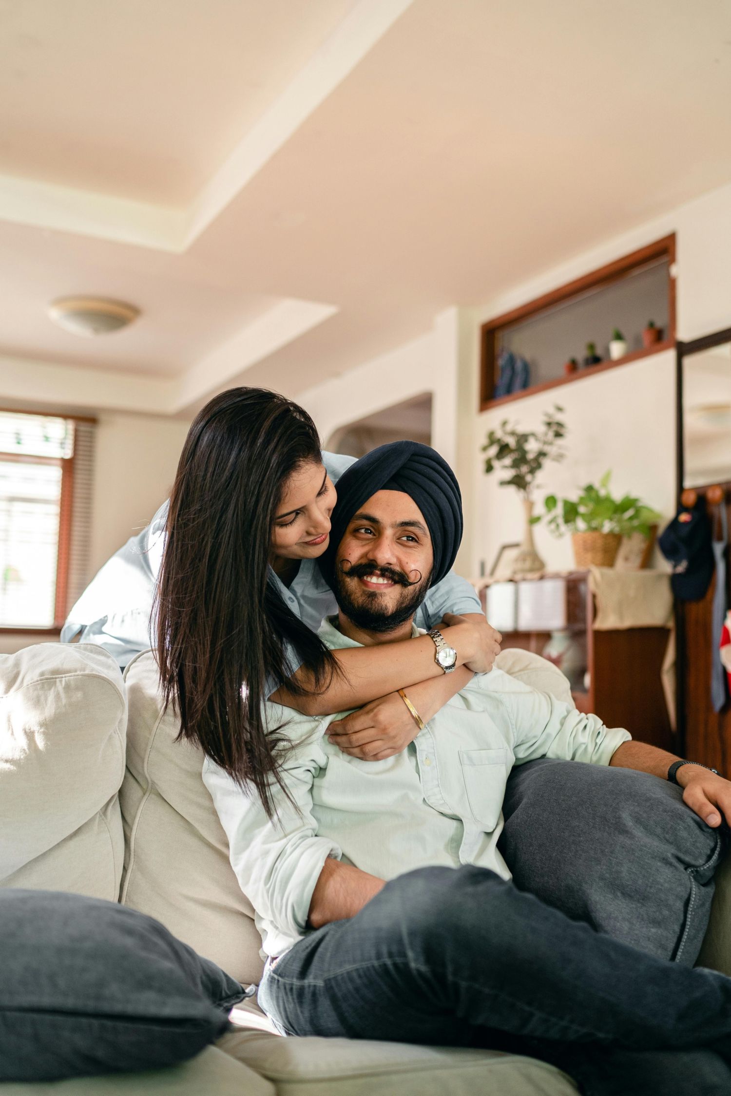 Cheerful young Indian female in casual wear hugging smiling Sikh husband in turban sitting relaxed on couch with legs crossed at home