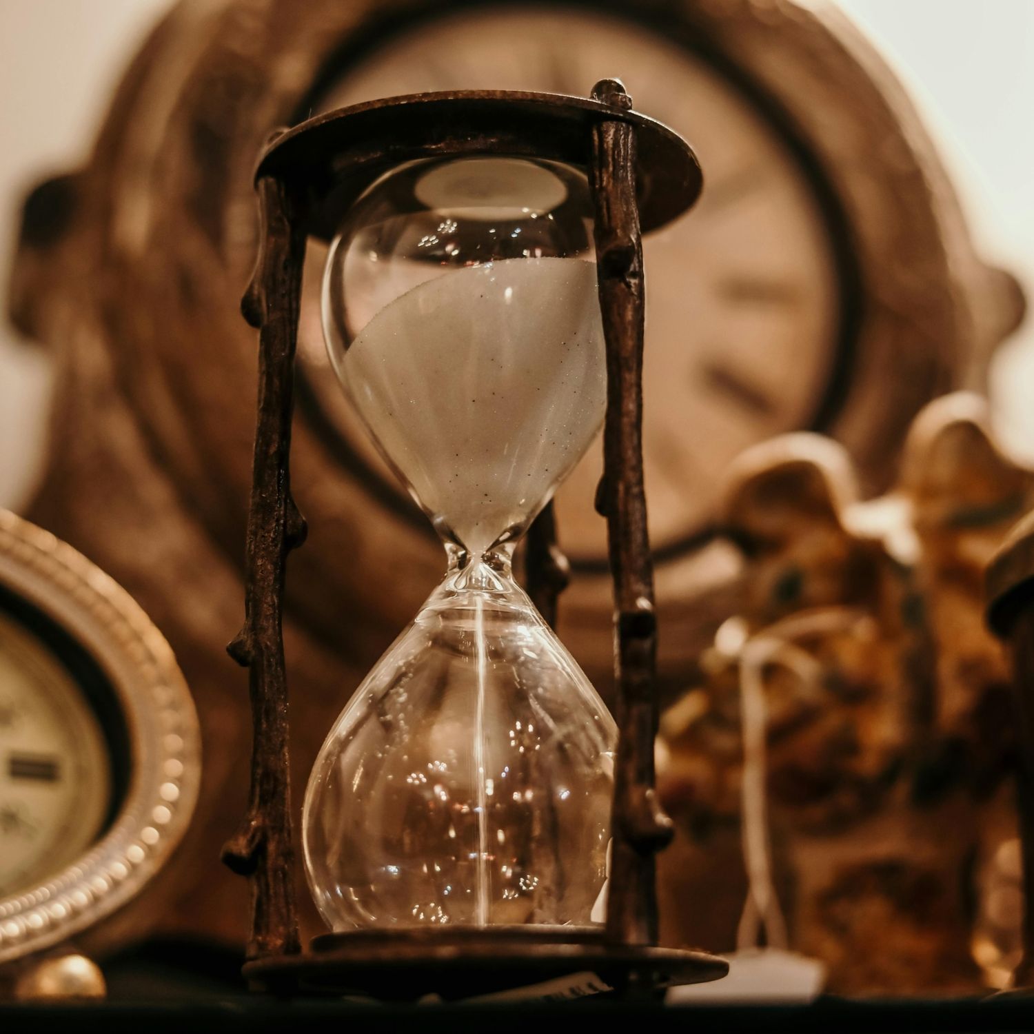 Close-up of a vintage hourglass surrounded by antique clocks in warm lighting.